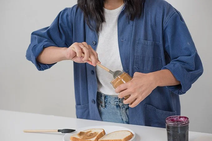 Person spreading peanut butter on a slice of bread with a spoon, on a white table, wearing a blue jacket and white t-shirt.