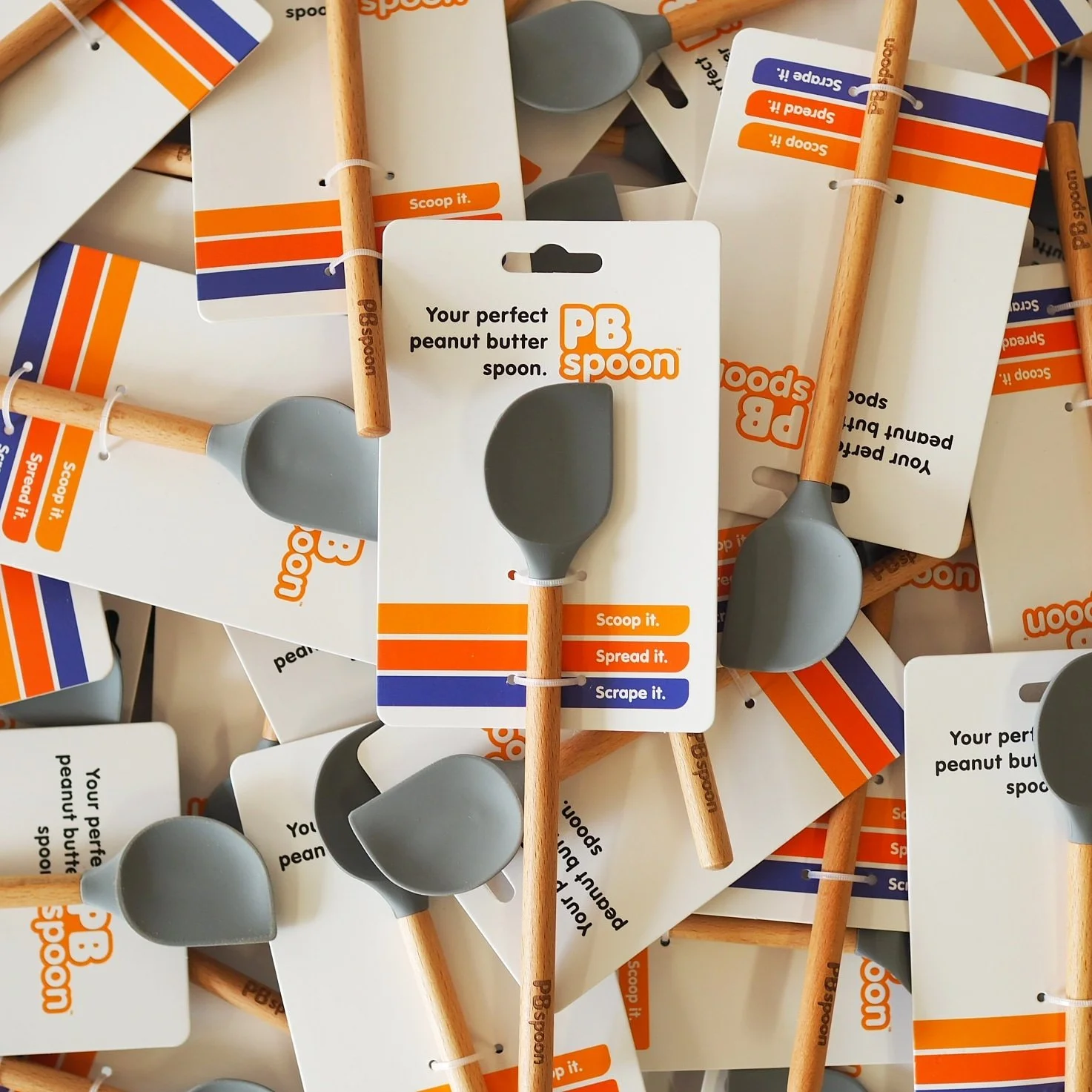 Pile of peanut butter spoons with wooden handles and gray silicone heads, attached to white cards with the PB Spoon logo and instructions to scoop, spread, and scrape peanut butter.