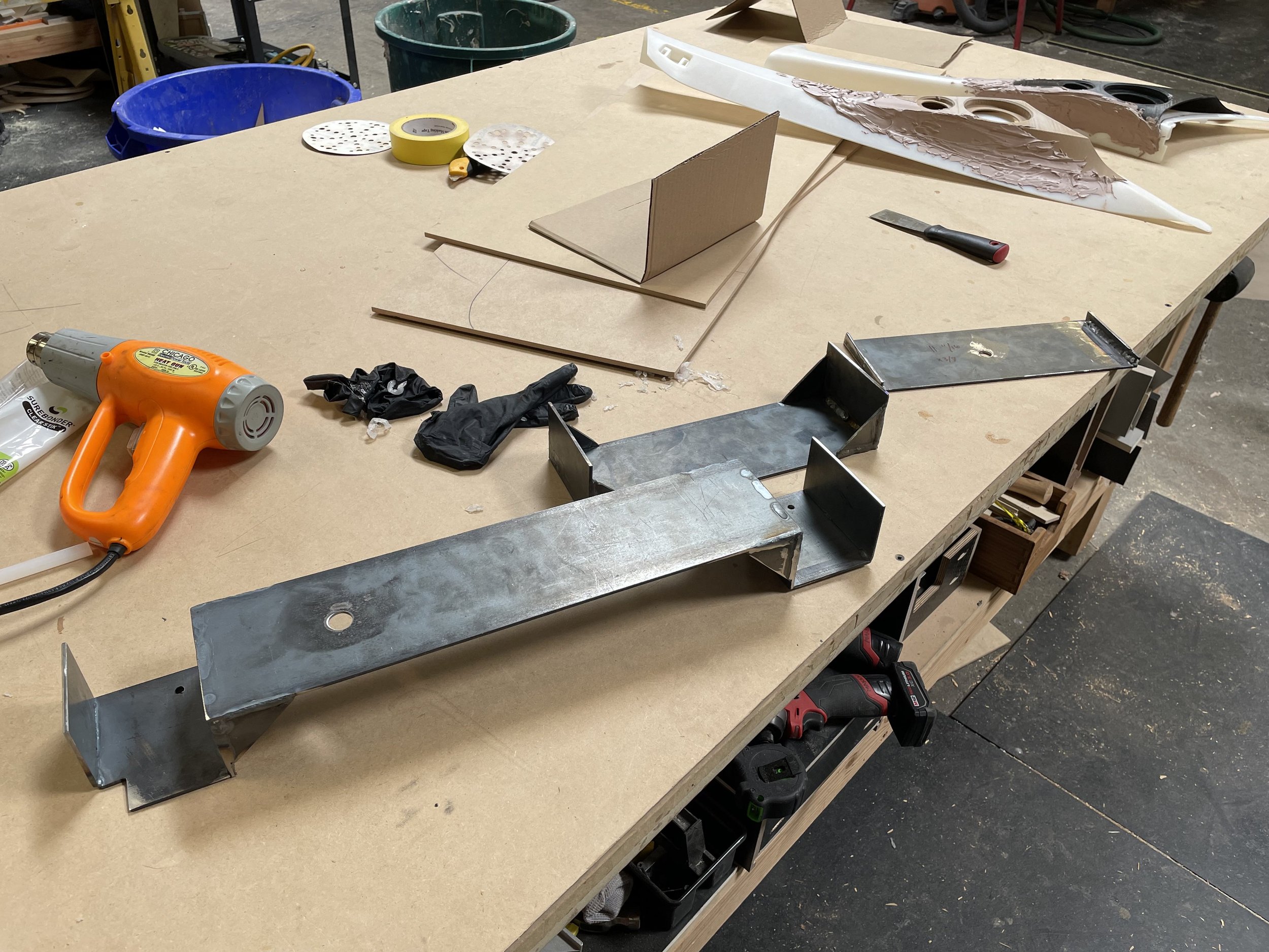 Work table in a workshop with various tools and materials, including metal parts, a heat gun, black gloves, and cardboard models, with a workbench in the background.