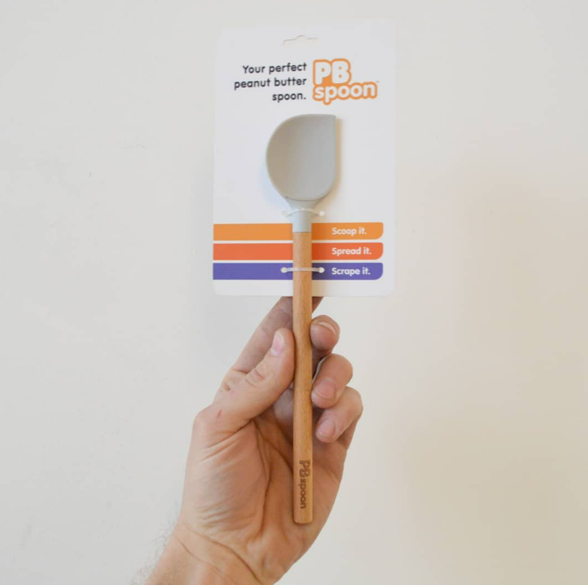 A hand holding a small peanut butter spoon with a cardboard backing. The spoon is labeled 'PB spoon' and has a silicone part. The cardboard background has instructions to scoop, spread, and scrape with colored bars.