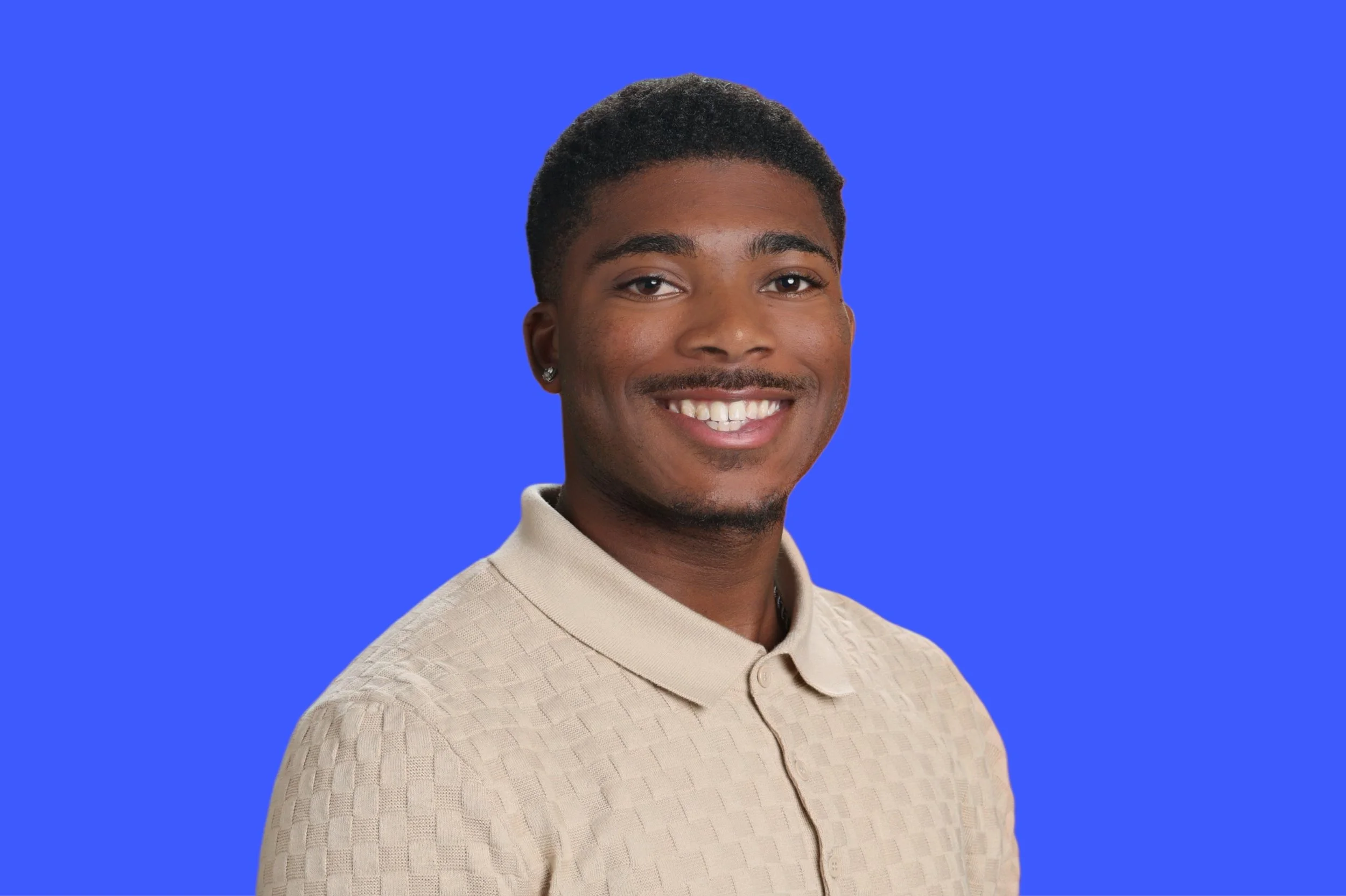 Professional portrait of Andrew McElvy smiling in a beige textured jacket against a clean blue background, representing a creative and professional presence within marketing, innovation, and brand development.