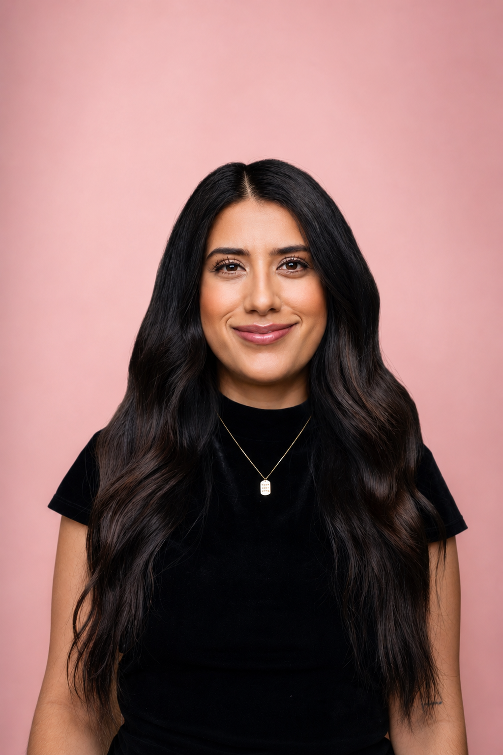 A woman with long, wavy dark hair smiling at the camera, wearing a black top and a gold necklace with a rectangular pendant, against a pink background.