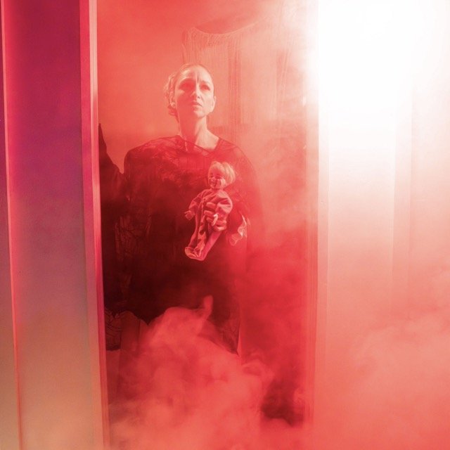 A woman taking a mirror selfie with a smartphone, holding a doll, surrounded by red smoke or fog.