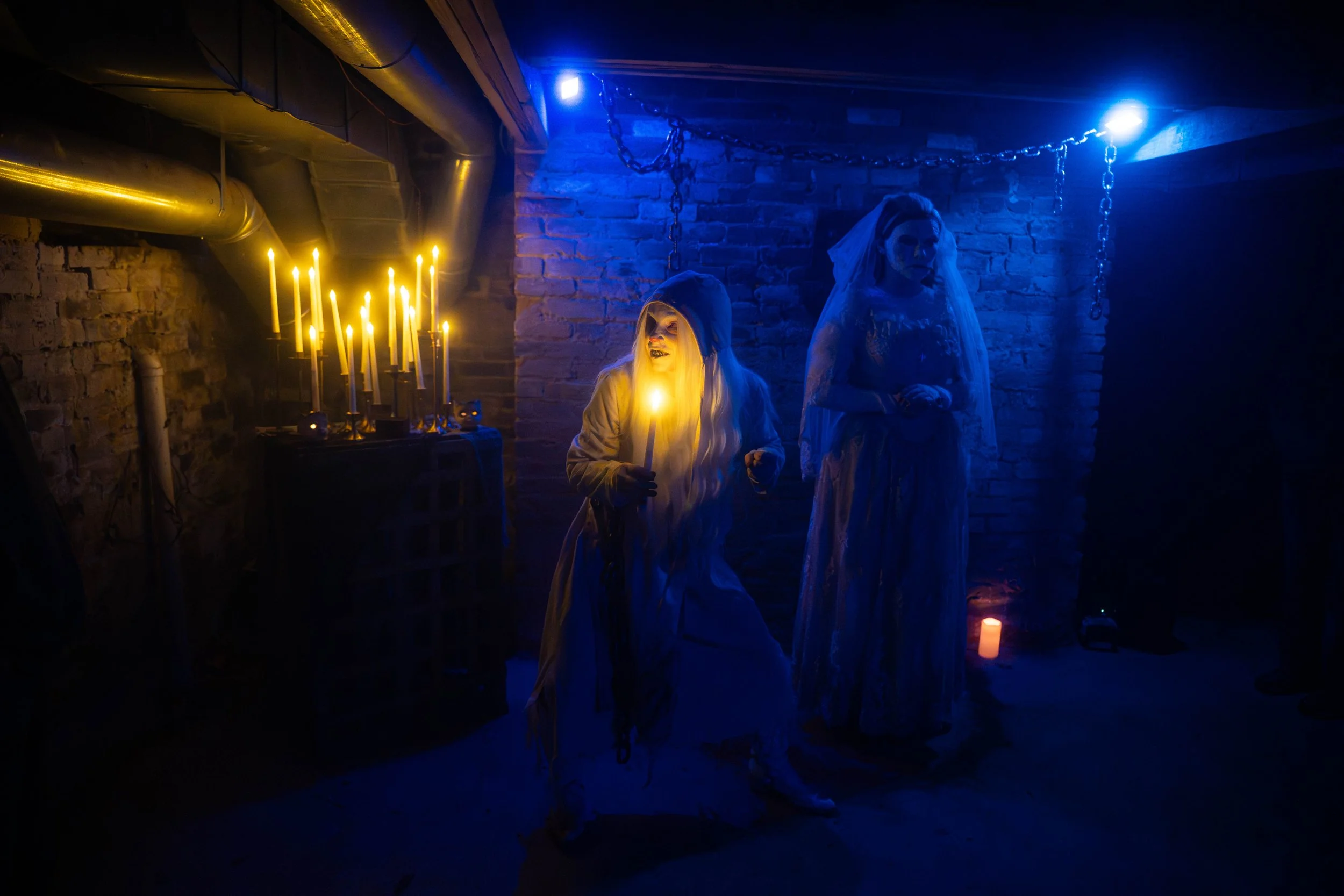 A dimly lit basement with exposed brick walls and chains hanging from the ceiling. Two ghostly figurines, draped in white fabric, stand among flickering candles, creating an eerie atmosphere.