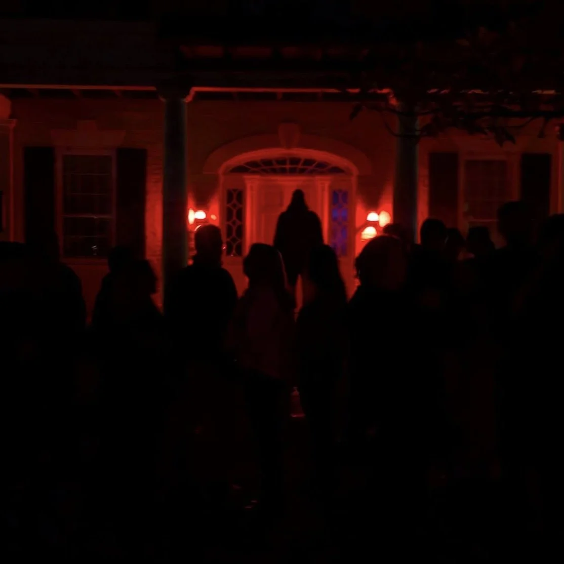 Silhouettes of people standing outside a house at night with red lighting, with a woman standing on the porch.