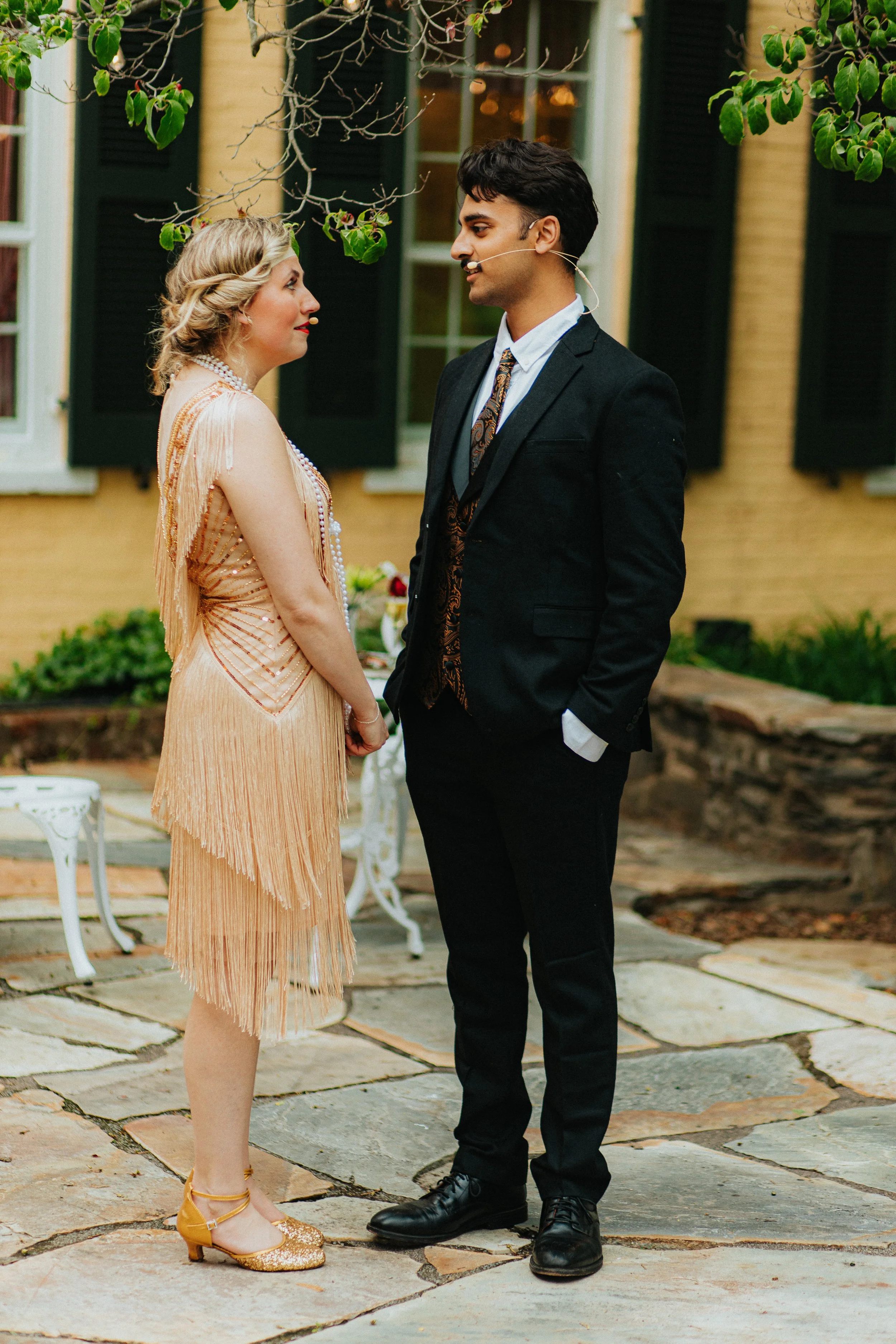A man and woman dressed in vintage attire facing each other in a garden outdoor setting, with a yellow building with green shutters in the background.