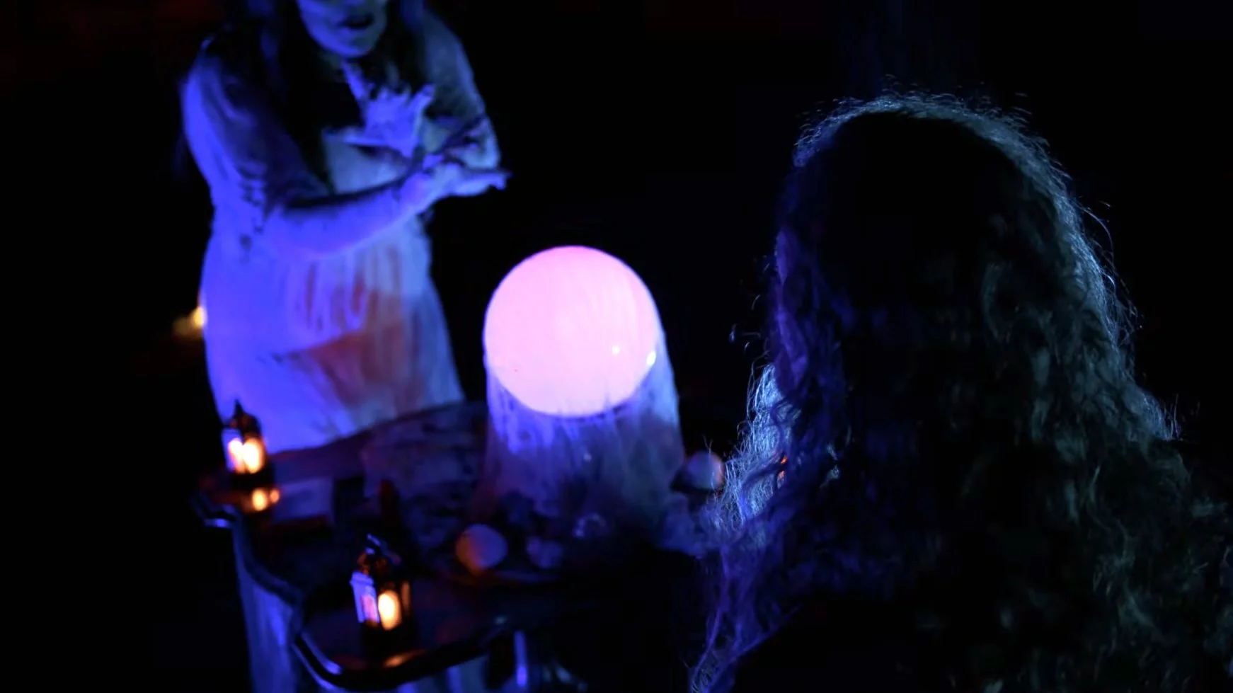 A woman with curly hair sitting at a table with a glowing purple orb and small candles, engaging in a tarot card reading.
