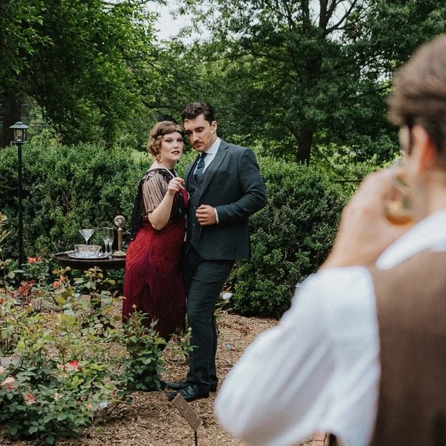 People at an outdoor garden party, with two individuals in vintage-style clothing posing and a third person in the foreground taking a photo.