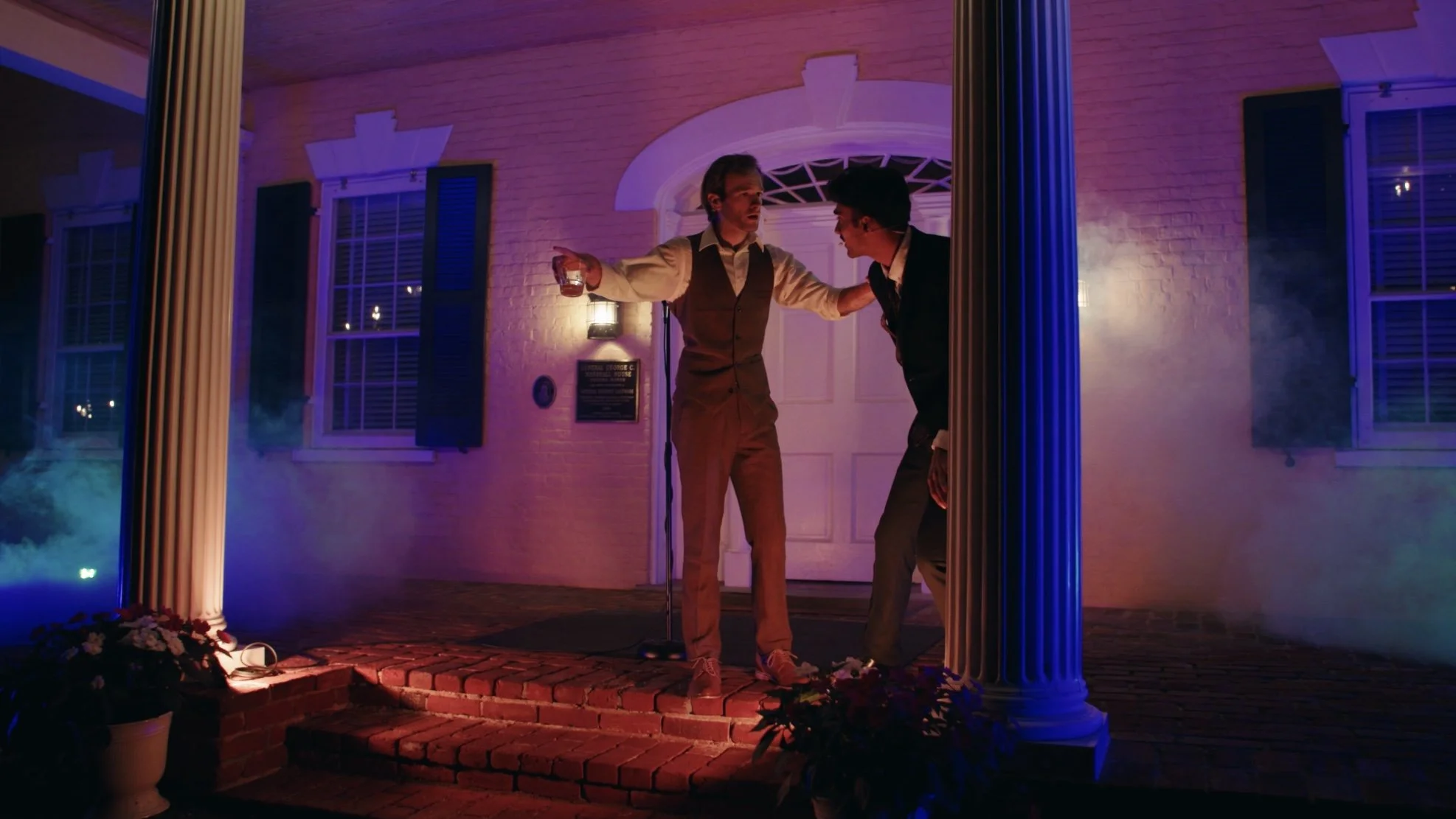 Two men dressed in formal attire standing on porch at night, one holding a drink and gesturing, the other leaning forward with hand on knee, illuminated by multicolored lights.