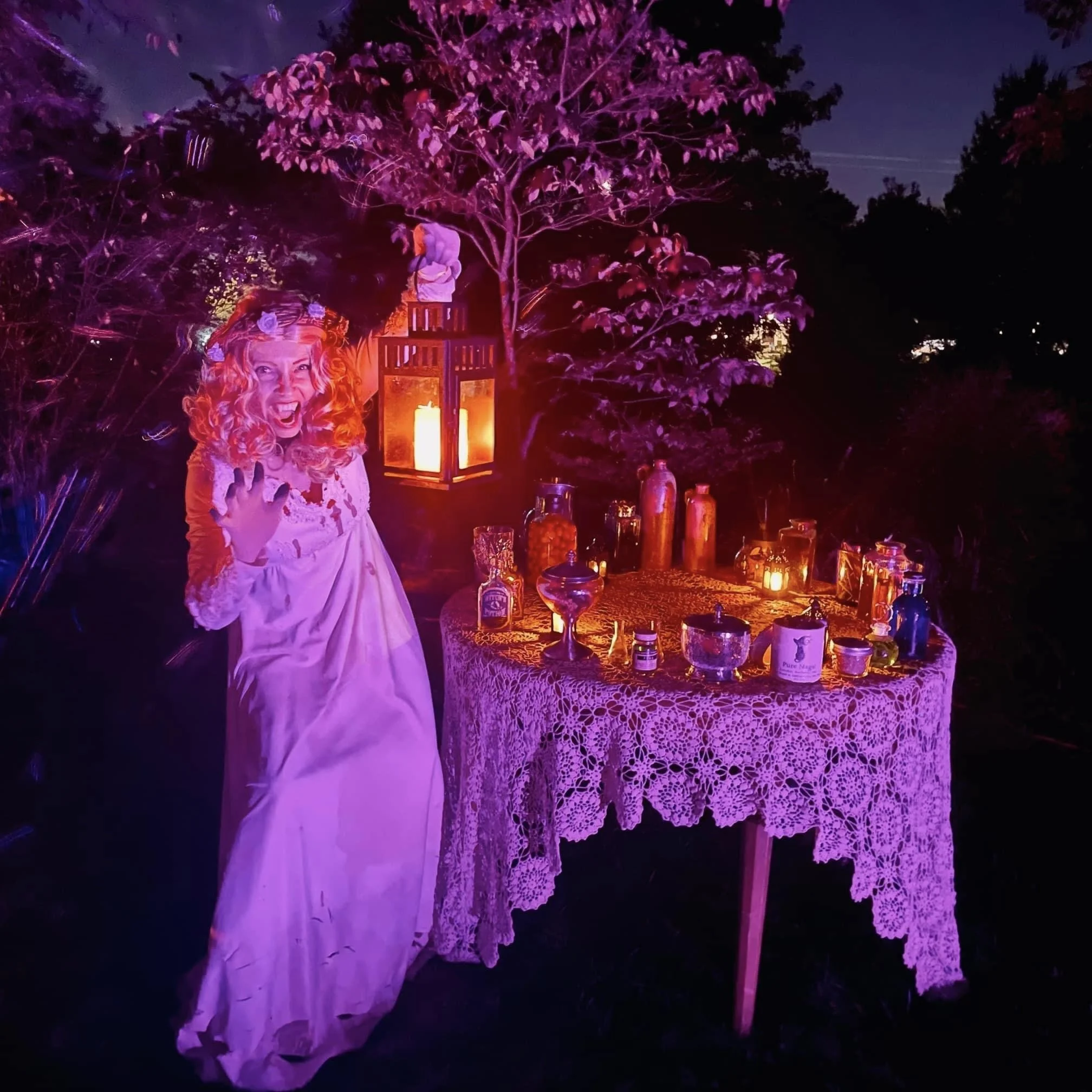 At night, a decorative scene with a person in a white dress and long, curly red hair, standing under a tree illuminated with purple and pink lighting. There are candles and various bottles on a lace-covered table.