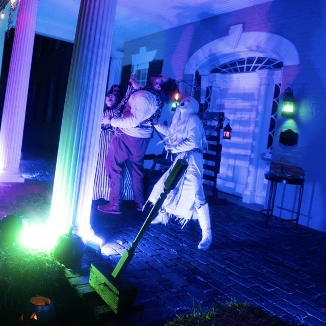 People in costumes dancing near a white column illuminated with colorful lights outside a house during a Halloween celebration.