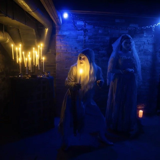 Two women dressed as witches or ghosts in a dark, spooky room with blue and yellow lighting, holding candles near a candelabrum, with a brick wall and chains hanging from the ceiling in the background.