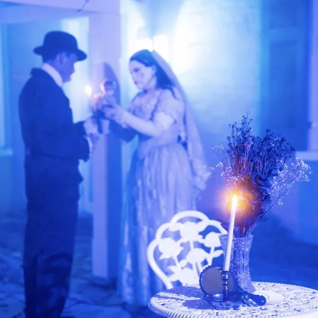 A vintage-themed wedding reception with a bride in a lace dress and veil and a groom in a dark suit and bowler hat, exchanging rings, lit by candlelight with a table decorated with flowers and a sparking candle.