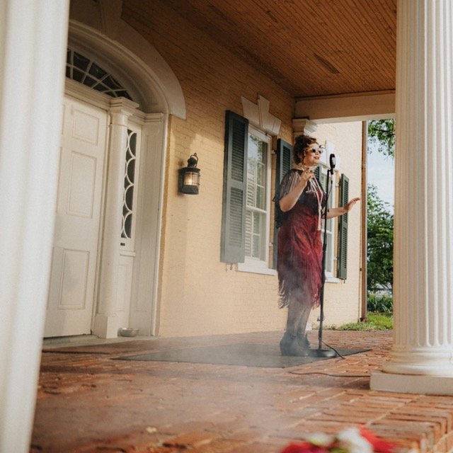Woman in vintage dress and sunglasses speaking into a microphone on front porch of a house with columns, brick floor, and open windows.