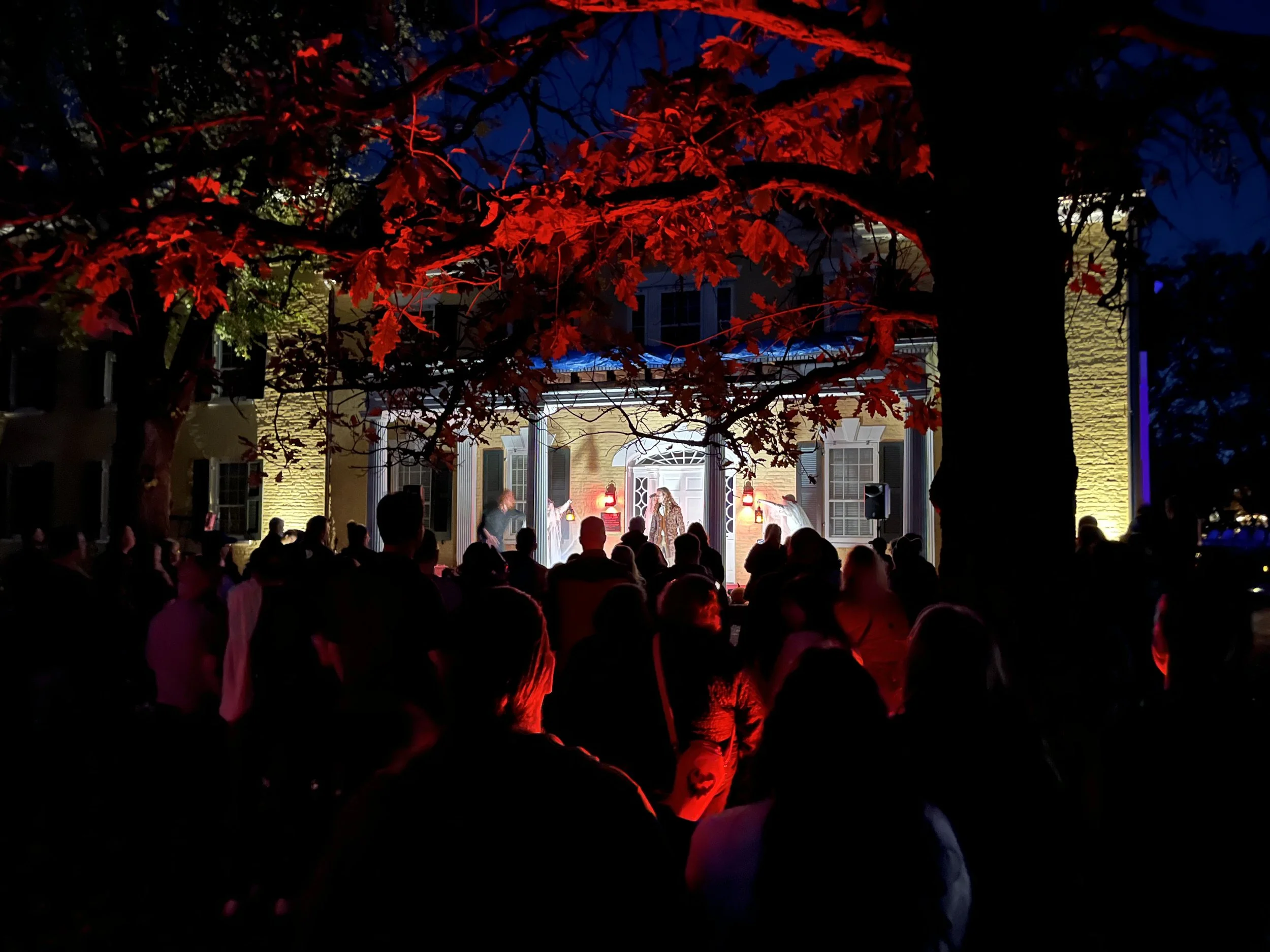 Nighttime outdoor concert with a crowd gathered in front of a stage illuminated with warm lights, framed by trees with red leaves, and a brick building with windows in the background.