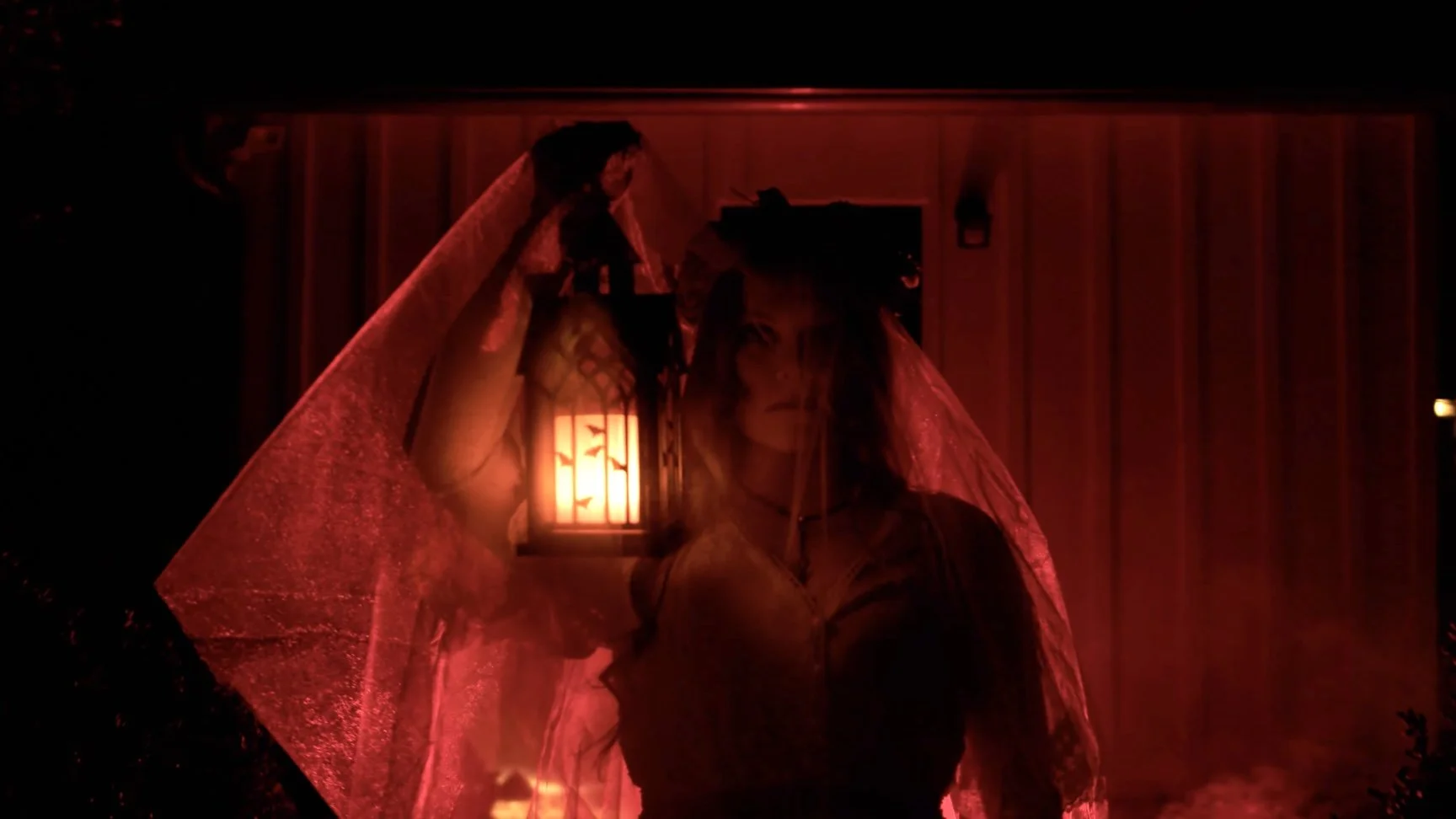 A person with long black hair and traditional Asian attire stands behind a red lace veil, illuminated by a lit lantern on a wooden wall background.