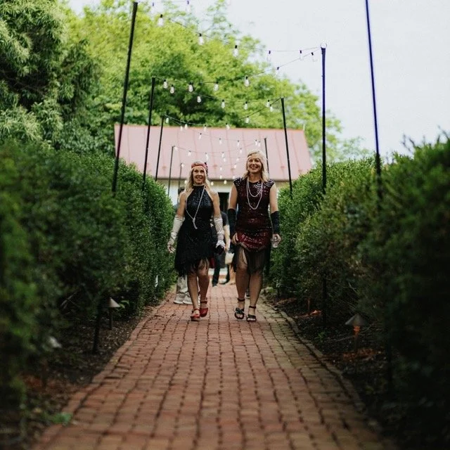 Two women dressed in 1920s flapper costumes walking on a brick pathway lined with bushes, with string lights overhead, in an outdoor garden setting.