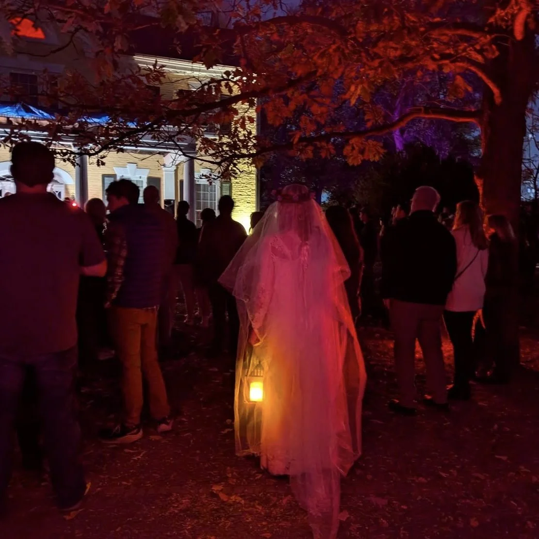 People gathered outdoors at night near a tree with red leaves, illuminated by colorful lights, with a woman dressed as a bride in a wedding gown and veil in the center.