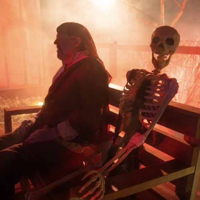 A person sitting on a bench next to a skeleton holding a scythe in a foggy, orange-lit scene.