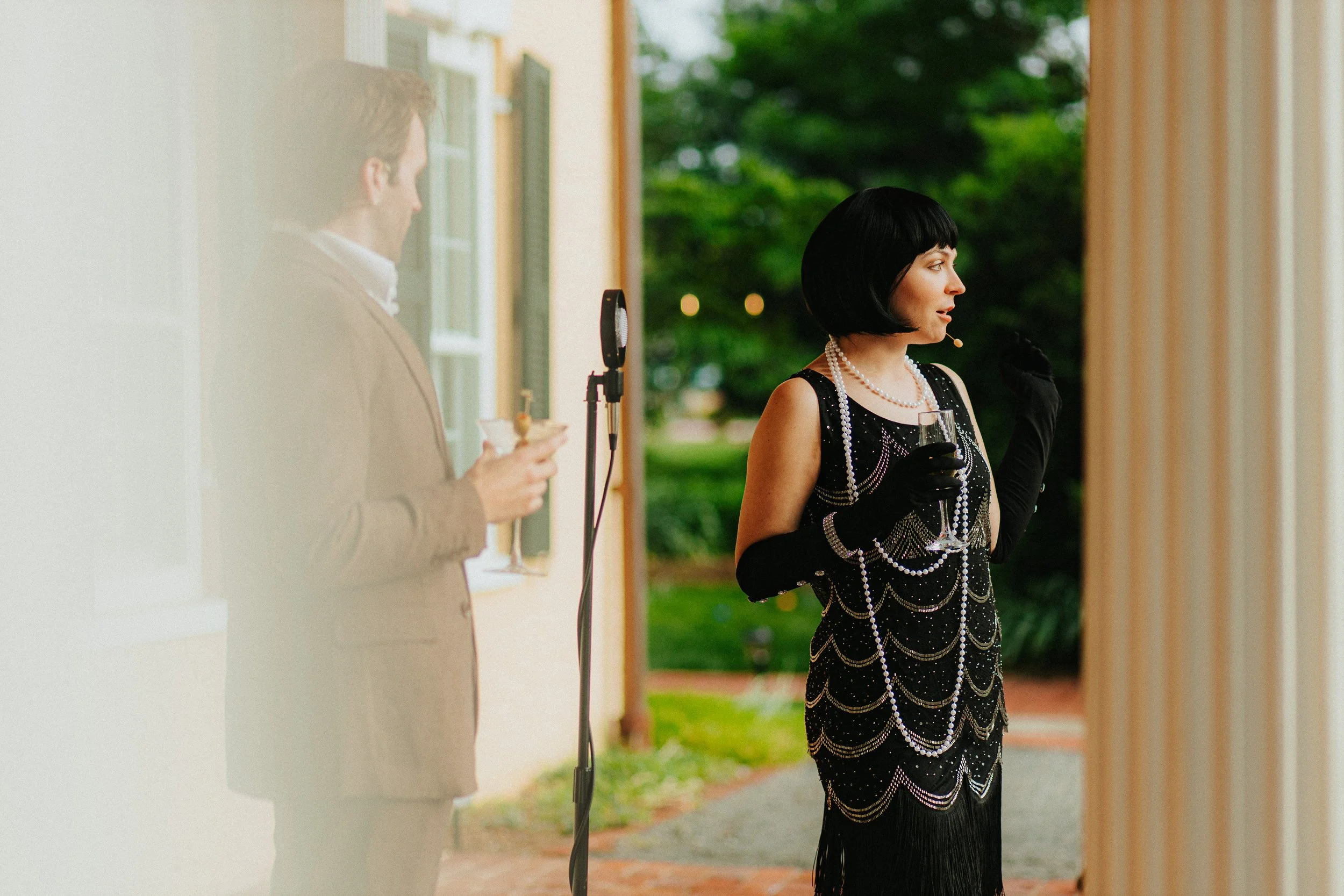 A woman dressed in a 1920s flapper style dress with pearl necklaces and black gloves, holding a glass, standing outside near a porch. A man in a beige suit is in the background, looking at his phone, near a microphone stand, with greenery and trees behind them.