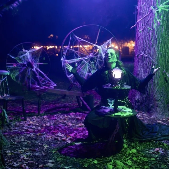 A woman dressed as a fortune teller or witch sits on the ground under a tree at night, with a purple and green lighting effect. She has her arms outstretched and is surrounded by large dreamcatchers with web-like patterns, illuminated by multicolored