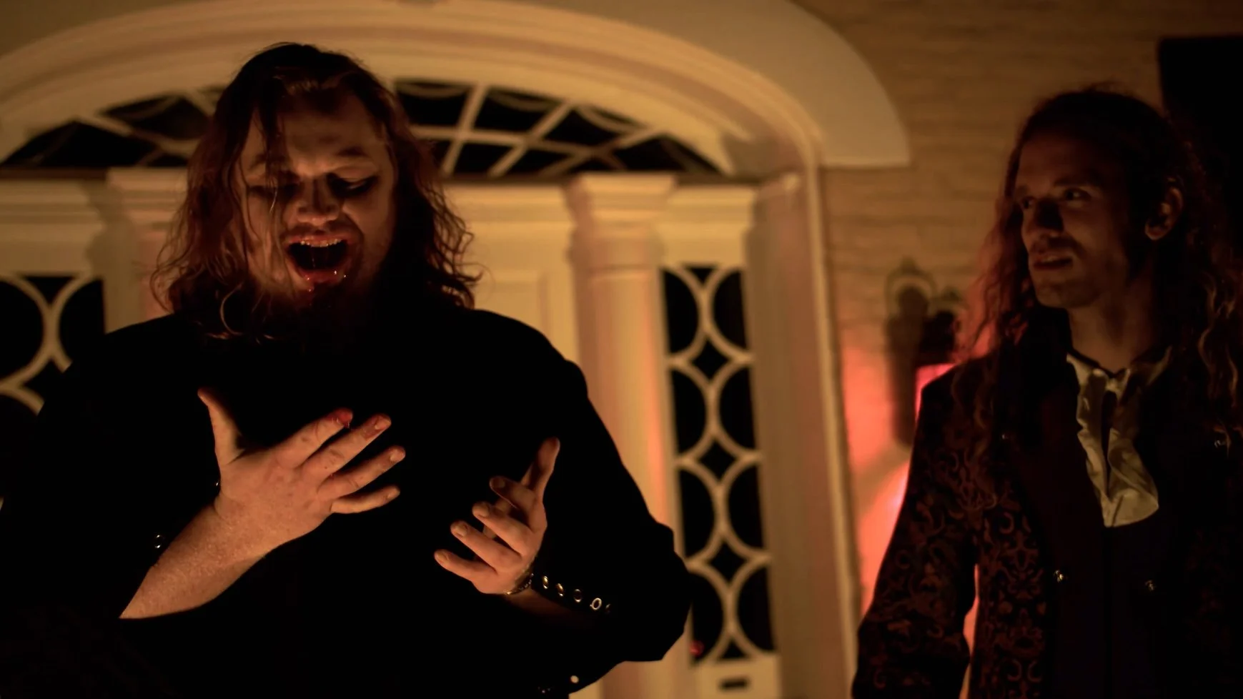 Two men with long hair at a party, one with his hand on his chest and the other looking at him, with warm lighting and decorative background.