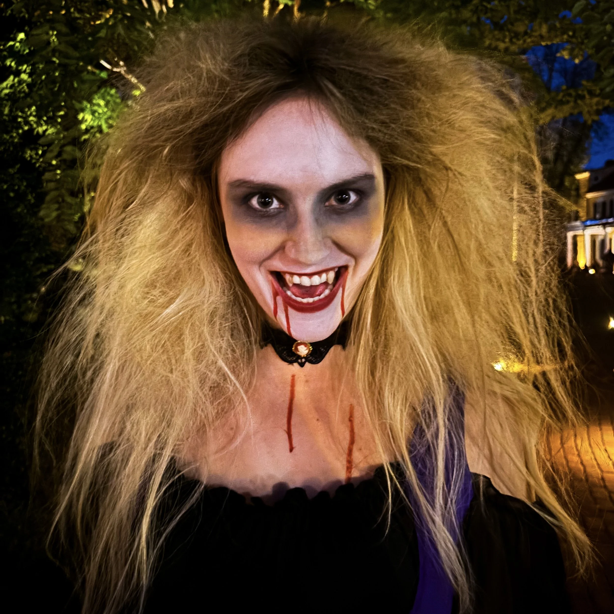 Person with wild, curly blonde hair, makeup, and vampire costume, smiling with an open mouth, night background with trees and houses.