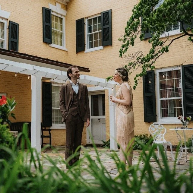 A man and woman dressed in formal attire standing in a garden patio, smiling and talking. The woman is holding a glass of champagne. The patio is part of a yellow house with black shutters and a white pergola overhead.