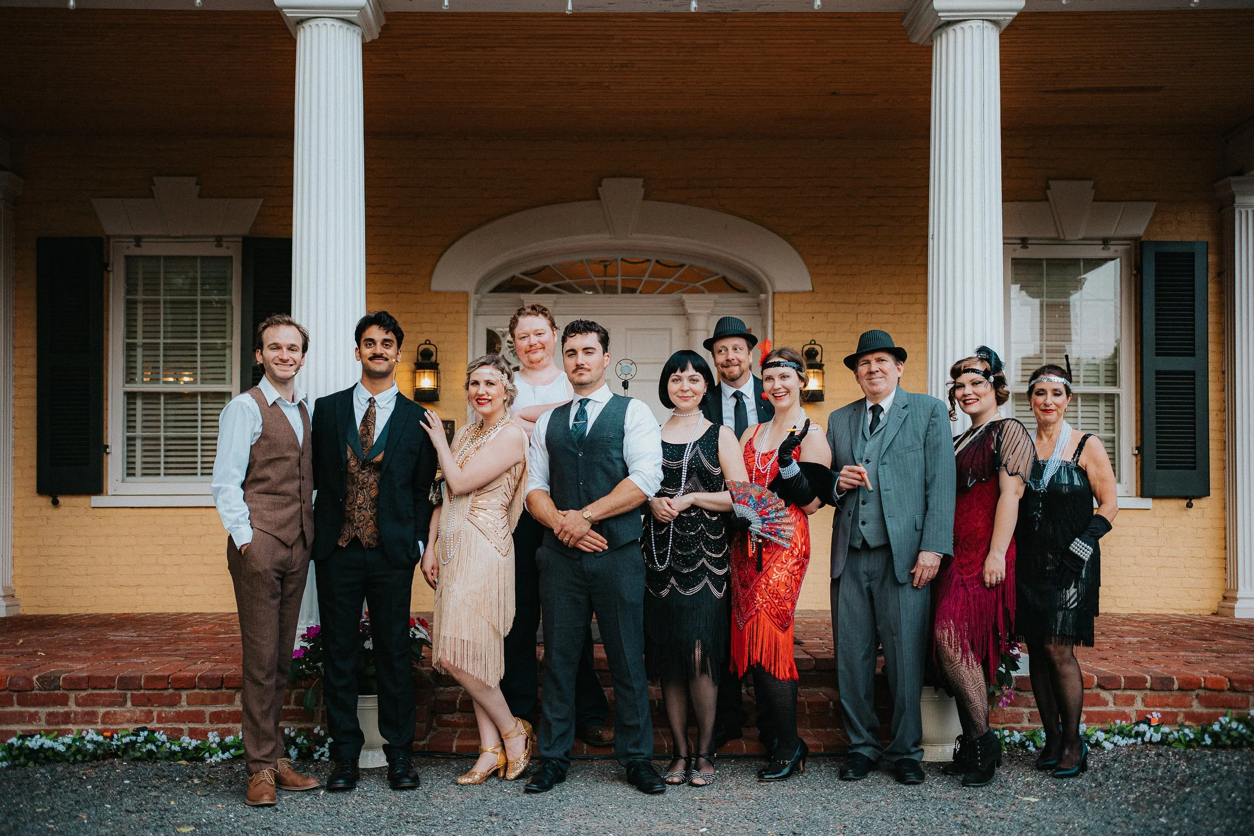 A group of eleven people dressed in vintage clothing standing on a brick porch in front of a yellow house with white columns. They are smiling and posing for a photo.