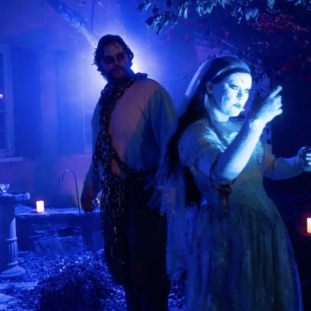 Two people in a dimly lit, outdoor setting with blue and purple lighting, surrounded by candles and foliage.