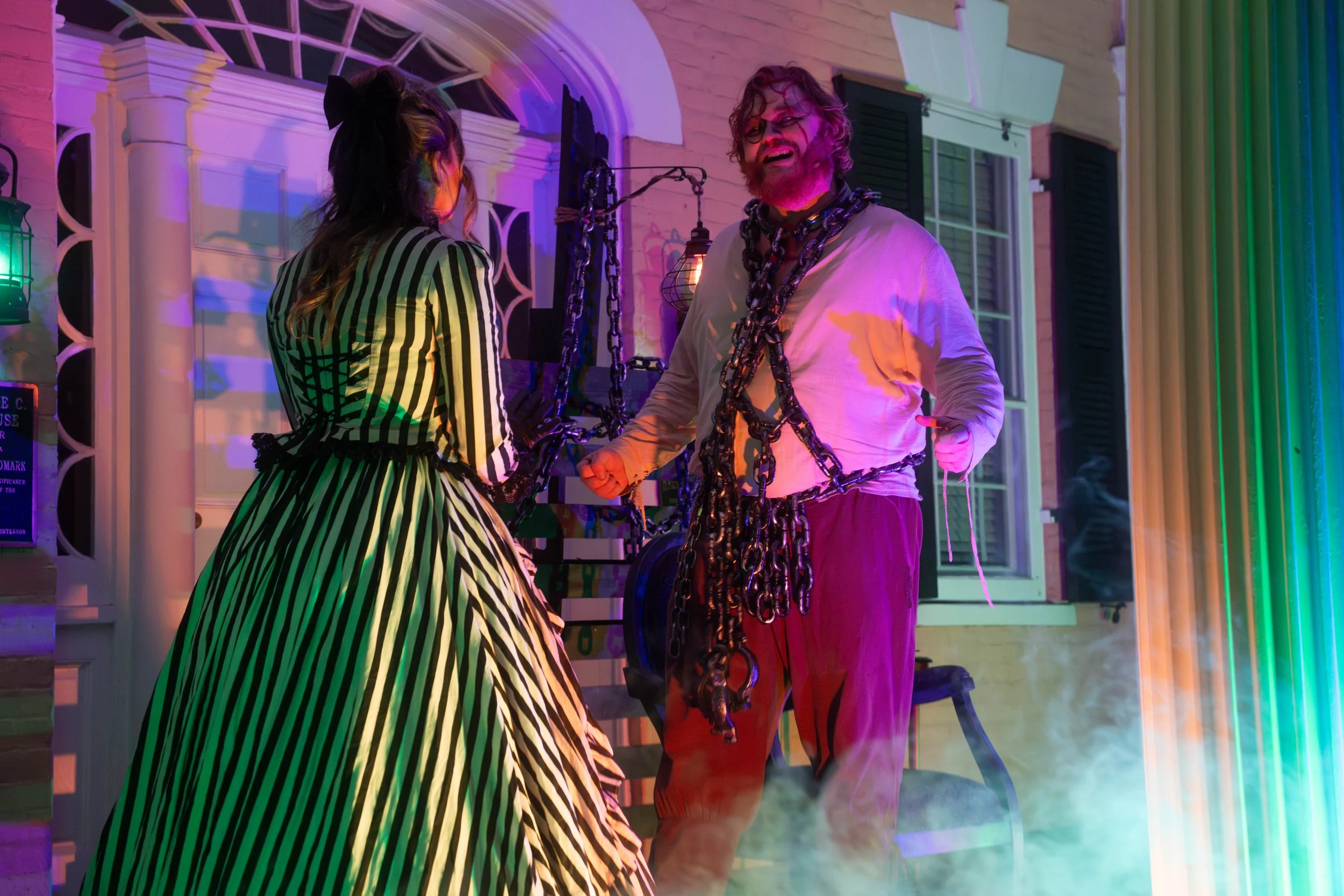 Two people are standing outside a house at night, illuminated by colorful lights. One person wears a black and white striped dress with a large black bow, and the other wears a white shirt and purple pants. The person in white has chains draped around their neck and shoulders. The scene has a spooky or theatrical vibe, with a background of a house with windows and greenery.