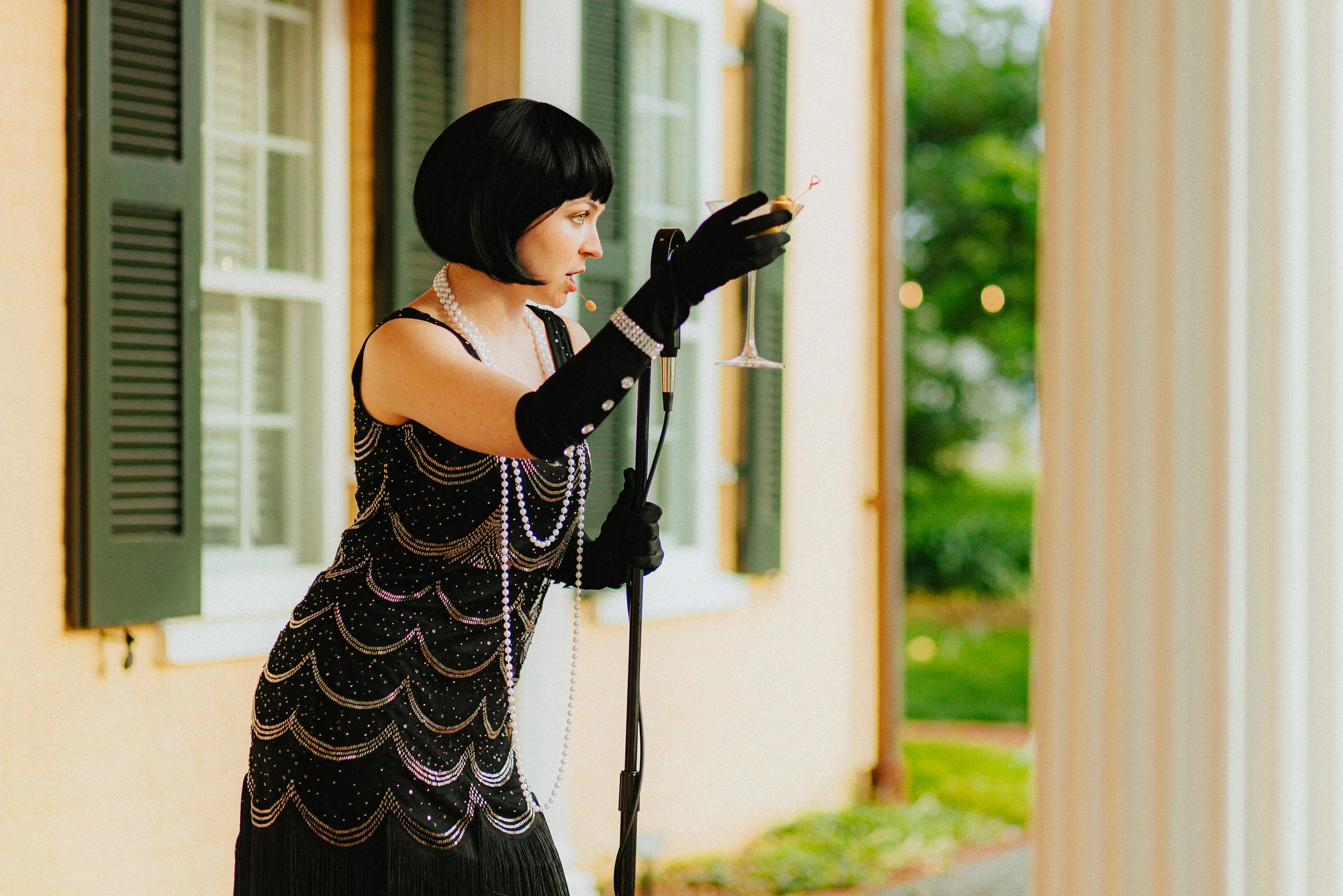 A woman dressed in a vintage beaded black dress with pearl accessories and long black gloves, holding a martini glass, standing in front of a porch with green shutters and white walls, with blurred green trees in the background.