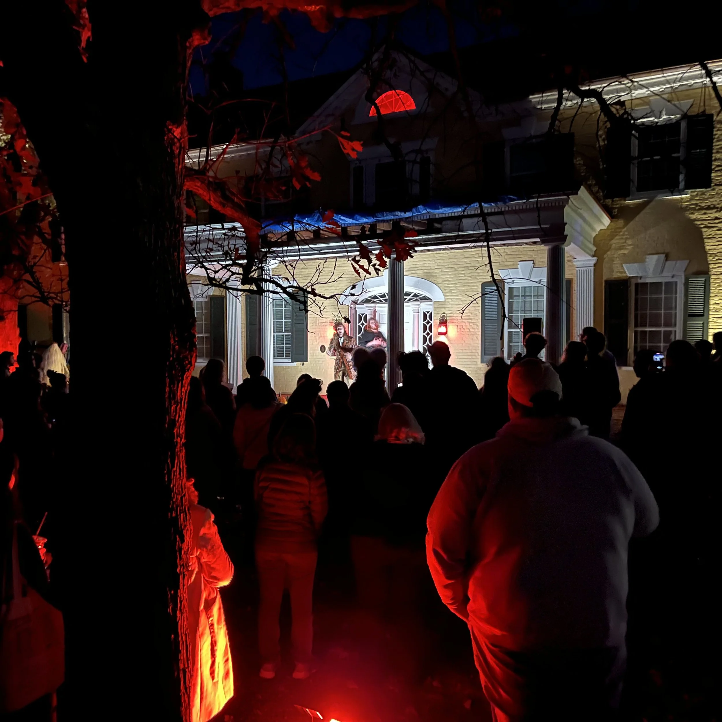 People gathered outside a house at night, illuminated by red lights, with some individuals engaged in conversation or activities near the front porch.