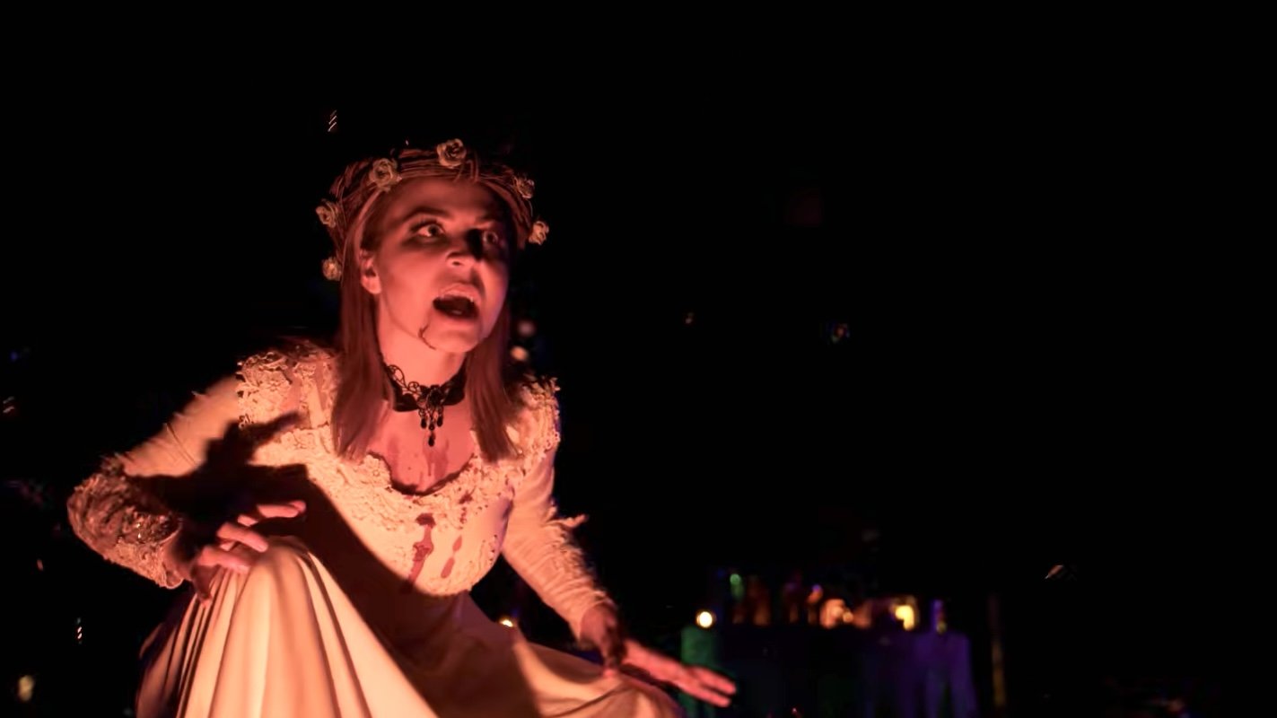 A person dressed in a costume with a lace top and a long skirt, wearing a crown of roses, making an expressive face with mouth open, in a dark setting with subtle colorful lights.