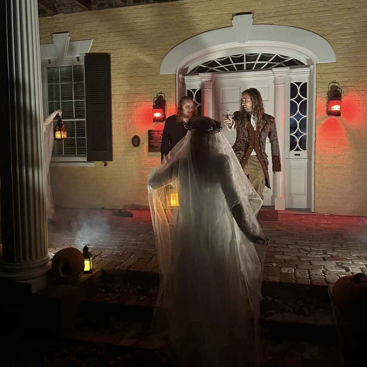 A person dressed as a bride in a wedding gown with a veil faces away from the camera, outdoors at night. Two men are standing in front of a house with a yellow brick wall, white door, and decorative columns, one of whom is holding a gun, and the othe