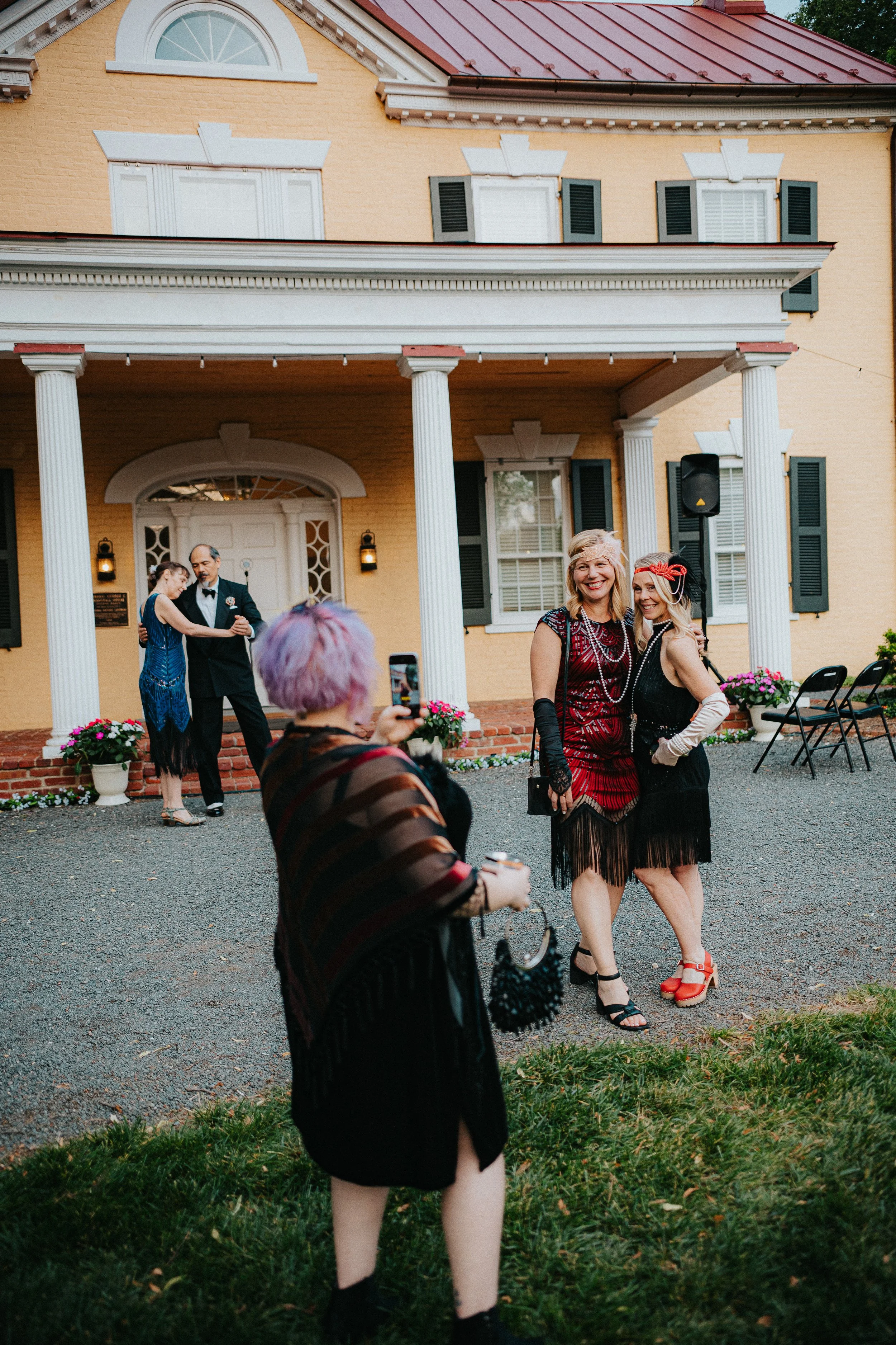 People dressed in vintage 1920s-style clothing at an outdoor event in front of a large yellow house with white columns. Two women are smiling and posing, while other guests dance in the background.