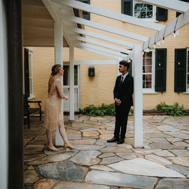 A man in a suit and a woman in a dress standing under a white pergola in a courtyard with a stone floor, yellow brick building with black shutters in the background.