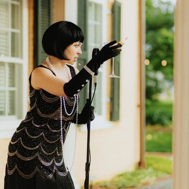 Woman dressed in 1920s style clothing, wearing long pearl necklaces, black gloves, and a short bob wig, holding a microphone stand and performing.