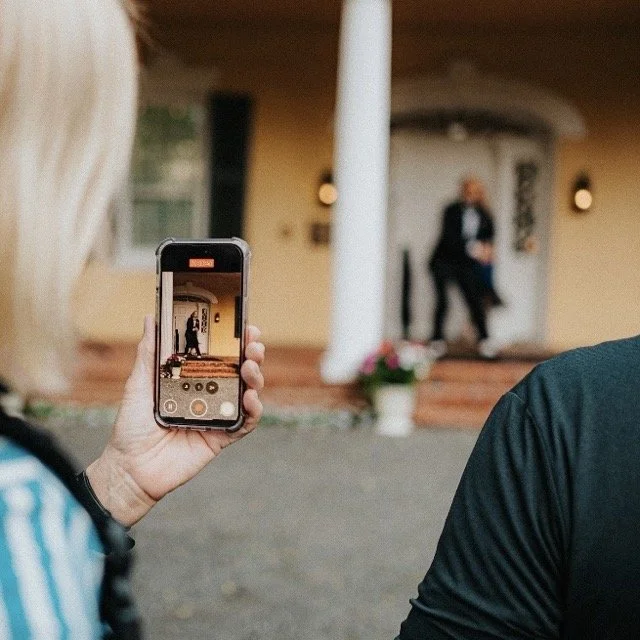 Person taking a photo of a couple dancing on a porch with a smartphone, with the couple slightly blurred in the background.