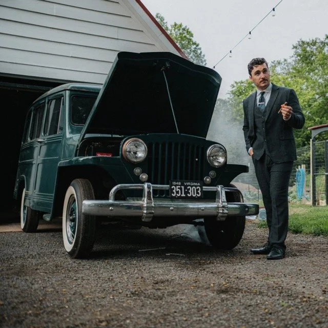 A man in a suit pointing, next to a vintage blue car with its hood open, in a backyard with trees and string lights.