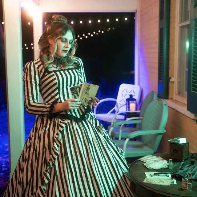 A woman with wavy hair wearing a black and white striped dress reading a book on a porch at night, with chairs, a lantern, and a table with papers and bottles outside.