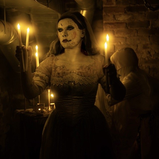 Woman dressed in ghoulish costume with painted face holding lit candles in a dimly lit room with brick wall background.