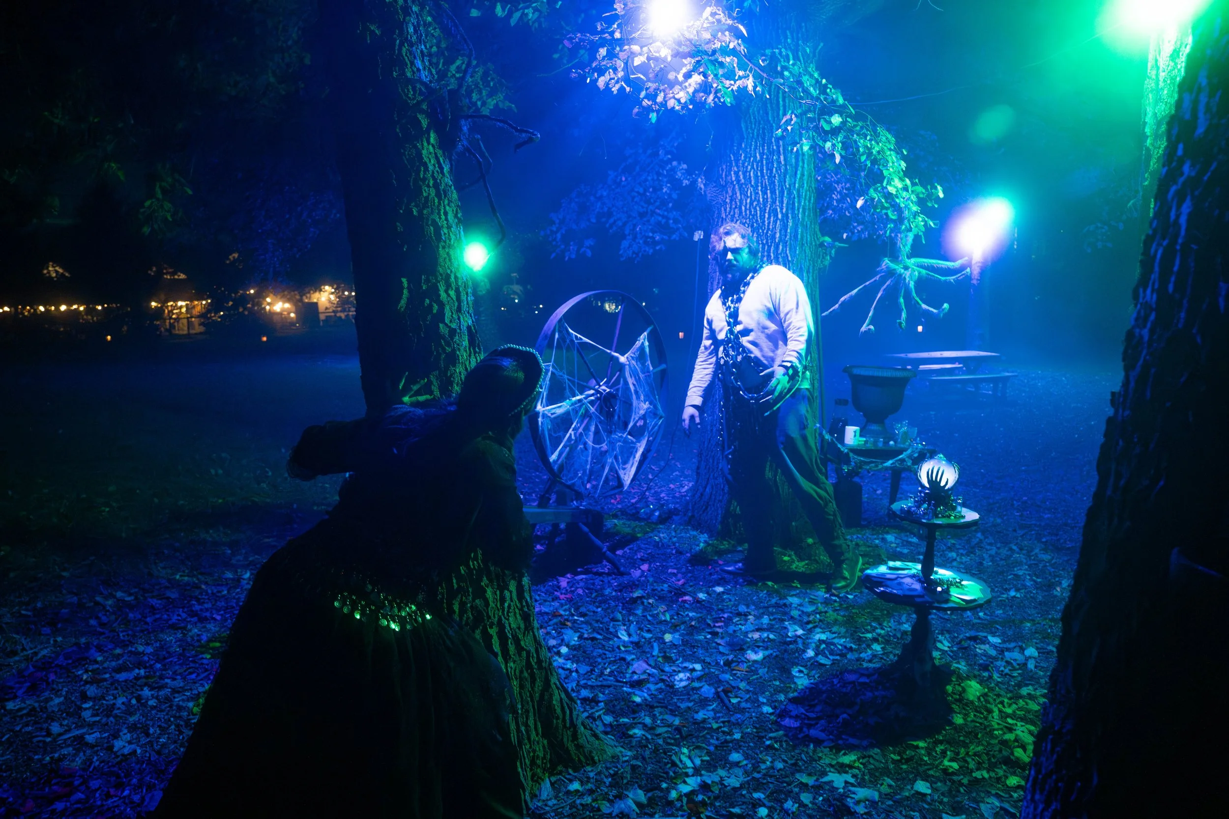 A nighttime outdoor scene with two people near large trees, illuminated by colorful blue, green, and purple lights. One person is sitting and the other is standing next to a table with art supplies, possibly creating art or engaging in a mystical activity. The background shows a row of small lights, suggesting a park or festival setting.
