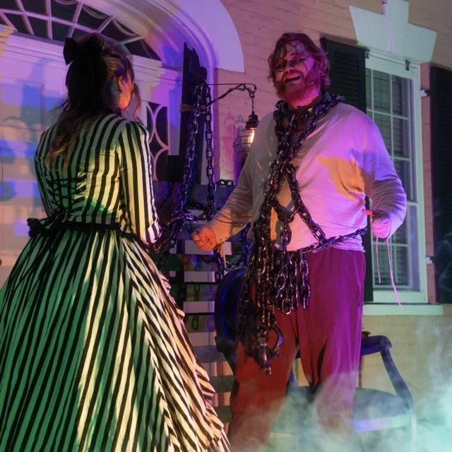 A man dressed as a scientist with chains around his neck and waist, holding a nerve in his hand, standing outside a house at night with colorful lighting and fog, talking to a woman in a vintage striped dress.