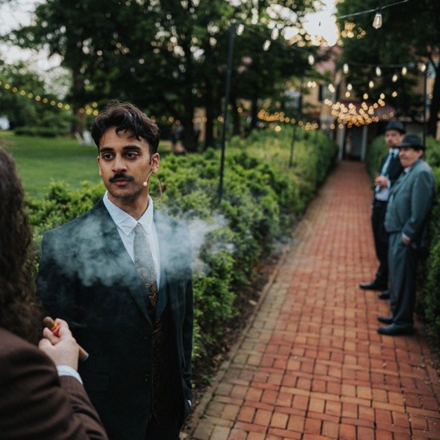 A man in a suit standing on a brick pathway during an outdoor event at dusk with string lights overhead, with other people in formal attire in the background.