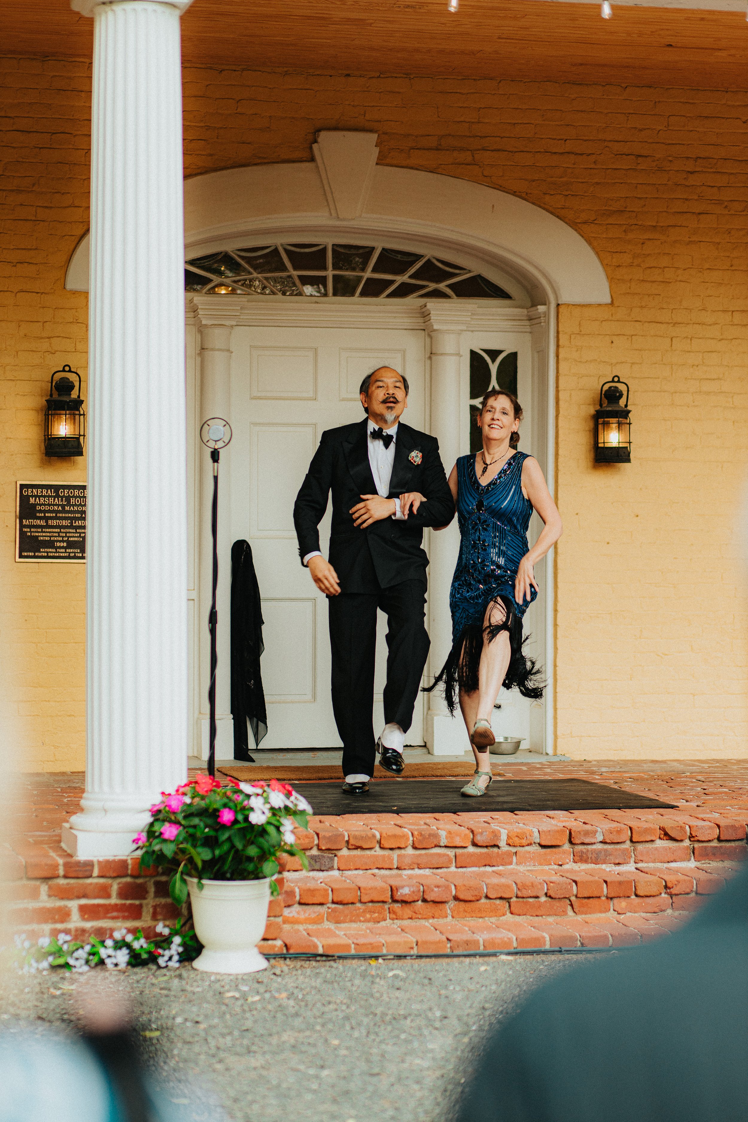 A man in a tuxedo and a woman in a fancy dress walk out of a building with a brick porch. There are two lantern-style lights on either side of the door and a flower pot with pink and white flowers on the steps.