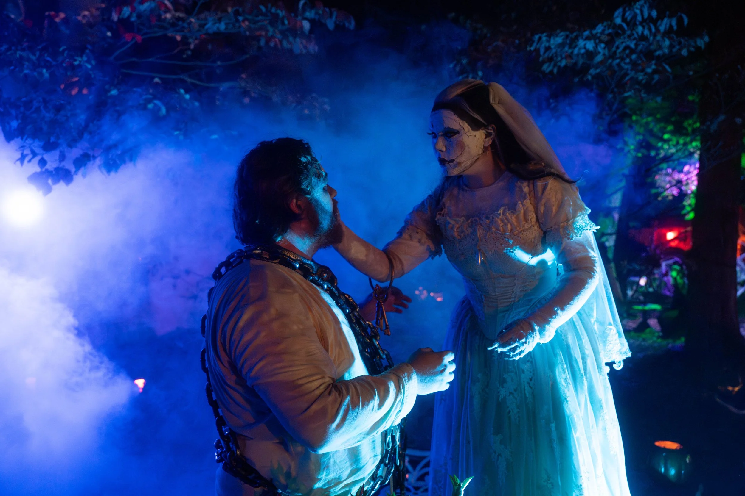 A person dressed as a bride with a skeletal face paint holding hands with a man in a suit at a nighttime outdoor event, surrounded by colorful lights and trees, with fog creating an eerie atmosphere.