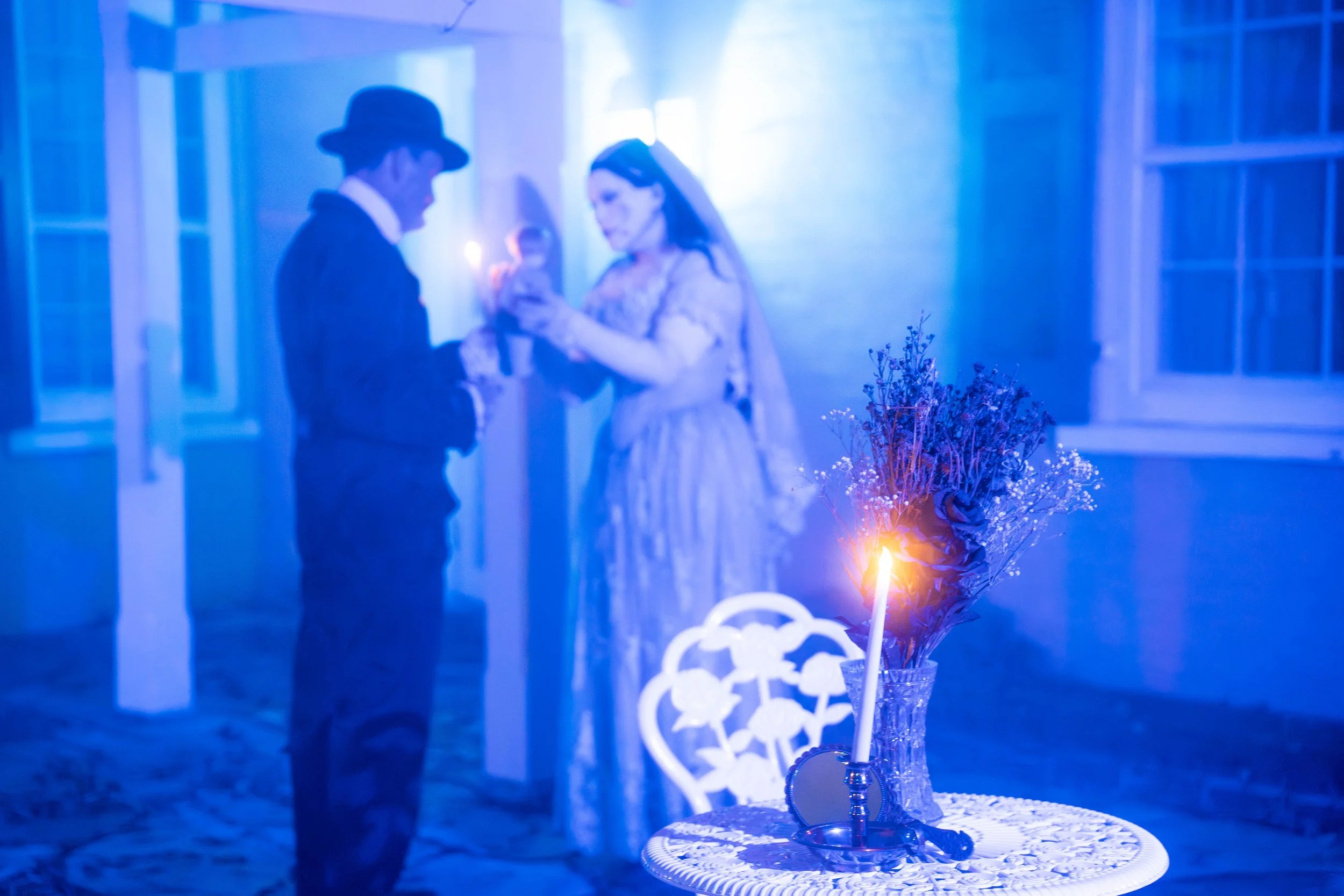 Couple dressed in formal attire, with the man wearing a hat and the woman in an elegant dress, standing close together in a blue-lit room, looking at a smartphone.