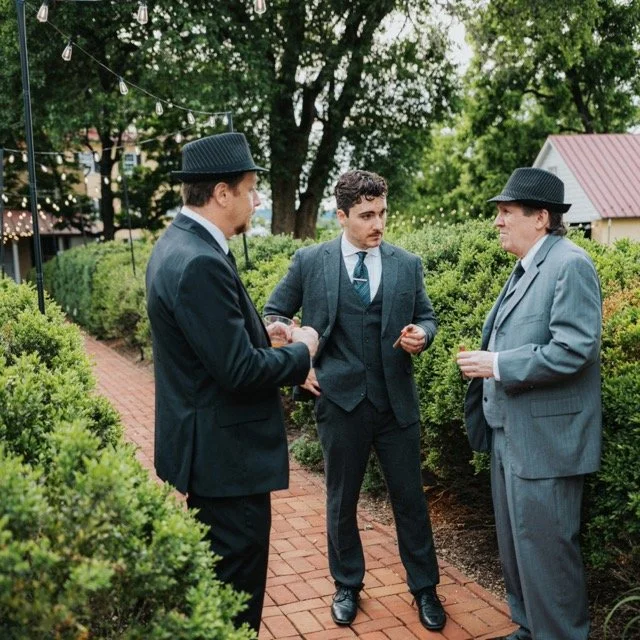 Three men in suits and hats standing on a brick pathway outdoors, engaging in conversation during an event.