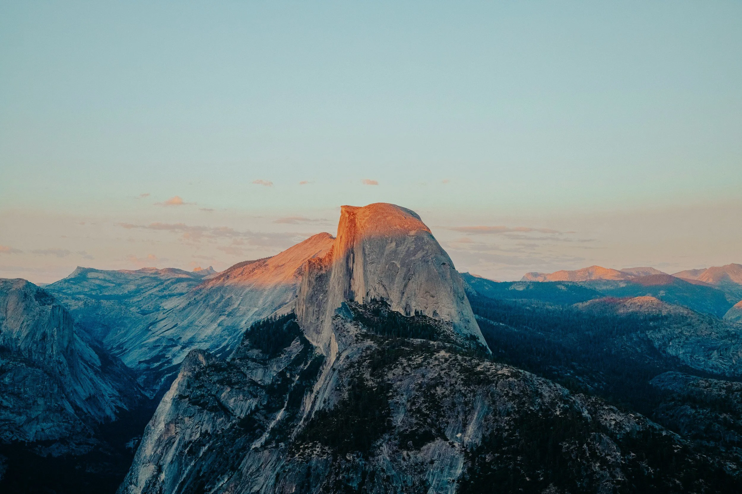 A mountain range with a prominent peak illuminated by sunlight, possibly during sunrise or sunset, with a sky featuring light pastel colors and a few scattered clouds.
